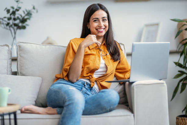 istock Pretty young woman working with her laptop while looking at camera sitting on couch at home. 2148624728