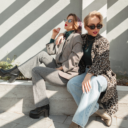 High fashion portrait of two stylish beautiful woman in trendy jackets and jeans sits and posing outdoor with sunlight.