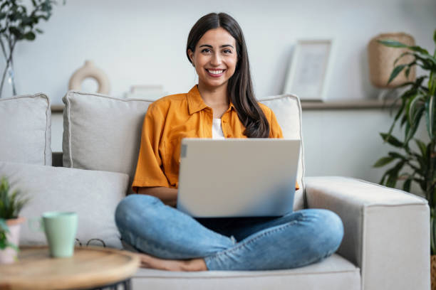 istock Smiling young woman working with her laptop while sitting on a couch at home 2148624943