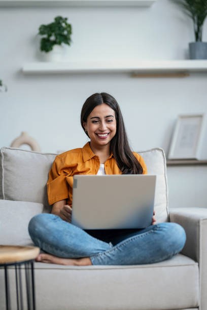 istock Smiling young woman working with her laptop while sitting on a couch at home 2148624948