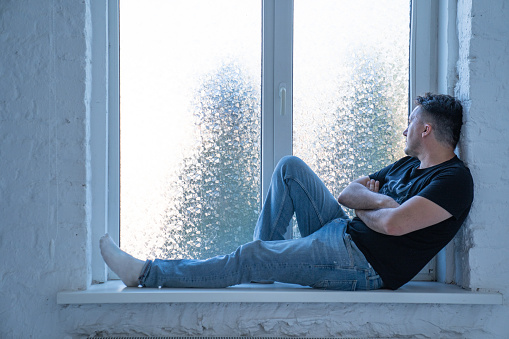 young handsome man with sad face, wearing jeans and t shirt is sitting on windowsill near the window, man looks through the window and thinks about life