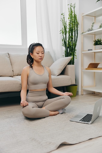 Asian woman in tracksuit sits with laptop in lotus position at home and meditates with eyes closed with a slight smile, mental health support lifestyle. High quality photo