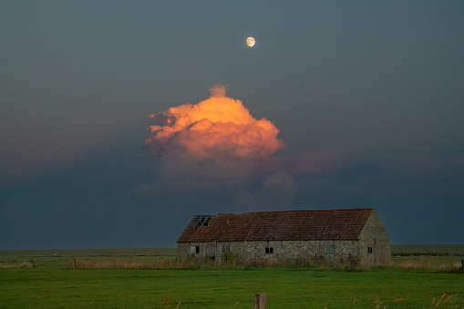 Golden cloud over old barn on Ameland, The Netherlands, 7/10/2014. Landscape with old barn and mysterious cloudy sky and moon. Idillic sunset with moon and golden cloud above old barn on Ameland