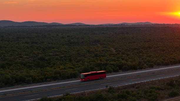 AERIAL: A lone red bus travels down the highway under a captivating crimson sunset, with a sprawling landscape around. Coach taking tourists on a road trip drives along a sunlit highway in Croatia.