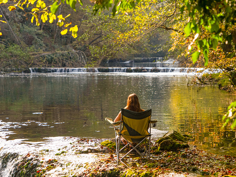 A young woman escaped from summer heat and is relaxing by the beautiful river. She sits on a camping chair and enjoys solitude and gentle sounds of free flowing water in pristine natural environment.