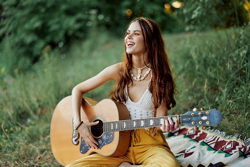 Young female hippie artist plays guitar and sings songs in eco-friendly clothing sitting on the ground outside in nature in the autumn looking out at the sunset. High quality photo