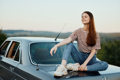 A young woman travels alone in her car on the roads in the countryside and relaxes sitting on the hood watching the sunset. High quality photo
