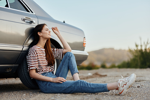 A young woman sits on the ground near her car on the side of the road and looks at the sunset. High quality photo