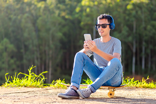 Young man seated on his longboard, joyfully enjoying music through wireless headphones, using music streaming service apps. He's outdoors, with many trees in the background