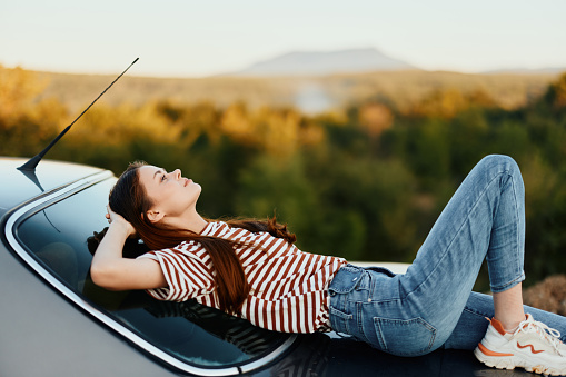 The woman driver stopped on the road and lay down on the car to rest and look at the beautiful landscape in a striped T-shirt and jeans. Complicated journey to nature. High quality photo