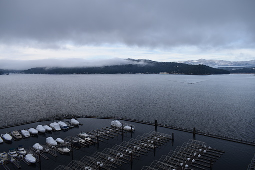 A wide angle view of Lake Coeur D'Alene in Idaho, USA. A lone boat is crossing the lake, heading towards the marina at the Coeur D'Alene Resort, leaving a wide reaching wake behind, and snow capped hills hiding behind low clouds in the background