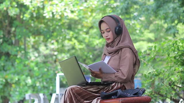 Beautiful asian woman wearing a hijab sits smiling operating a laptop while reading a book in the campus park. female student for education, technology and lifestyle concept
