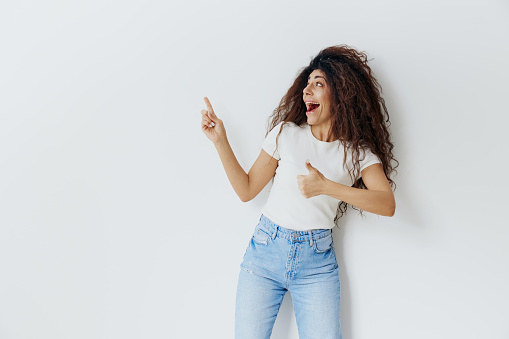 SEASONAL SALE OFFER. Cheerful, smiling curly Latin female stay near white wall, say Hey, Look at this, look up, show at free place copy space, cool ad for clothes sales. Studio shoot isolated