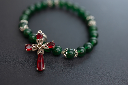 a beautifully crafted green beaded bracelet adorned with a silver and ruby cross pendant. The deep green beads are polished and evenly spaced, complementing the rich red of the rubies. The pendant, with its intricate design and embedded stones, hangs gracefully against the dark, matte background, highlighting the luster and luxury of the jewelry.