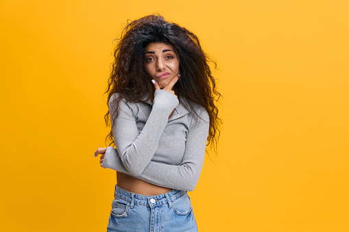 Portrait Of Pensive Latin Female Touching Chin And Looking At Camera with Doubt, Thoughtful Young Arab Woman Considering Interesting Offer While Standing Isolated Over Yellow Background, Copy Space