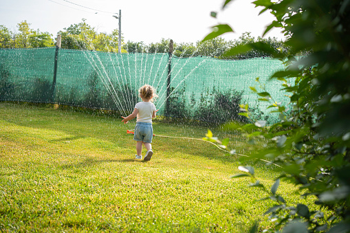 Cute little girl playing with water from garden hose sprinkler in a backyard.