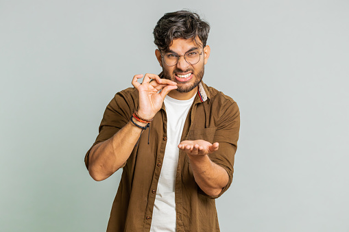 Need some more, please give me. Indian man showing a little bit gesture with sceptic smile showing minimum sign, measuring small size, begging help. Arabian guy isolated on gray studio background