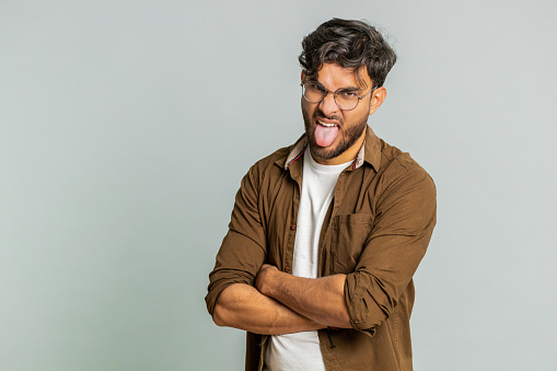 Playful Indian young man showing tongue making faces at camera, fooling around, joking, aping with silly face, teasing, bullying, abuse. Cheerful funny Arabian guy isolated on studio gray background