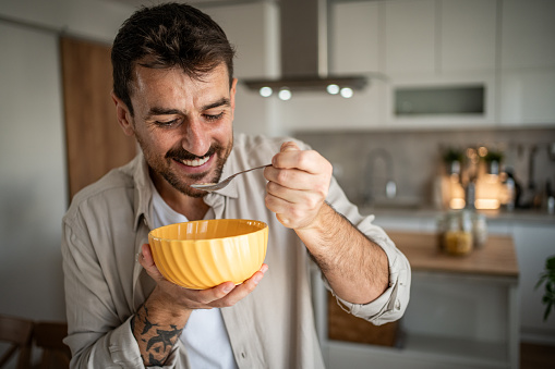 Photo shows a young man standing in his new apartment in the living room eating cornflakes for breakfast, morning routine