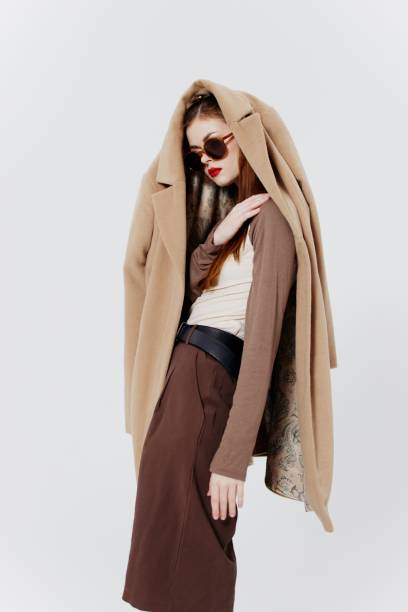 istock Stylish young model with a coat on her head poses with glasses in the studio 2148647113