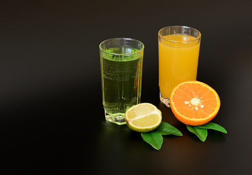 Two glasses with different citrus juices on a black background, next to half a lime and an orange with leaves. Close-up.