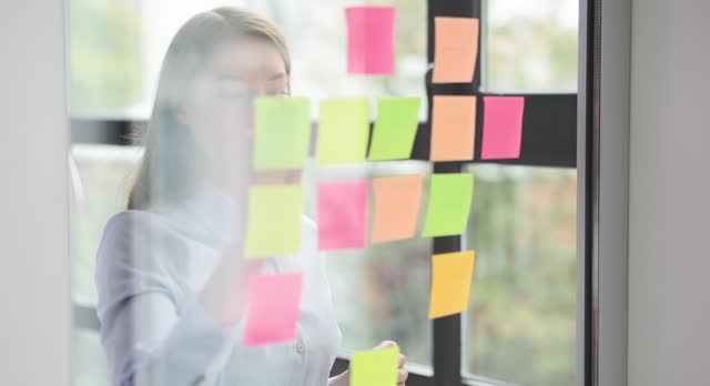 Beautiful female manager sticking colored note papers to a transparent glass wall in office