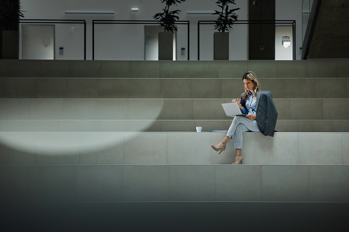 Young female entrepreneur talking to someone during a conference call over a computer while sitting on staircase in a hallway of an office building. Copy space.
