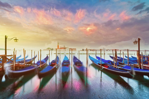 In the serene morning light of the Venetian Lagoon, a cluster of traditional gondolas are moored at a dock, evoking the essence of Italian culture and Venice's timeless beauty. Reflecting in the calm waters, they create a scene of tranquility and romance, capturing the allure of travel and the magic of this beloved destination.