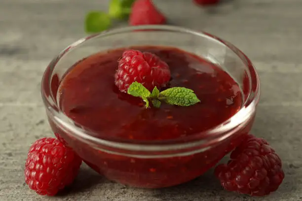 Photo of Glass bowl of raspberry jam with ingredients on gray textured background