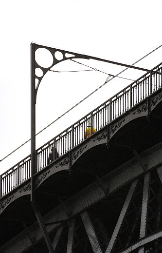 Man in yellow t-shirt and woman dressed in black with long hair, walking on the top of the Don Luis I steel bridge in Porto with details of its metal structure with ornaments under a cloudy white sky.