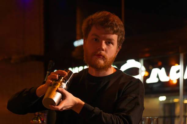 istock Professional bartender with a beard in a black sweatshirt holds a steel shaker behind the bar and looks at the camera. Bartender in a beautiful and atmospheric atmosphere in the bar. 2148674353
