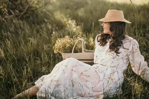 Artistic image of an adult woman sitting in the grass in a long dress and hat on her head.