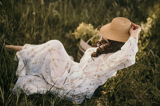 Artistic image of an adult woman sitting in the grass in a long dress and hat on her head.