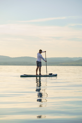 Joyful man is training SUP board on the lake at sunset. Stand up paddle boarding. Landscape in the background.