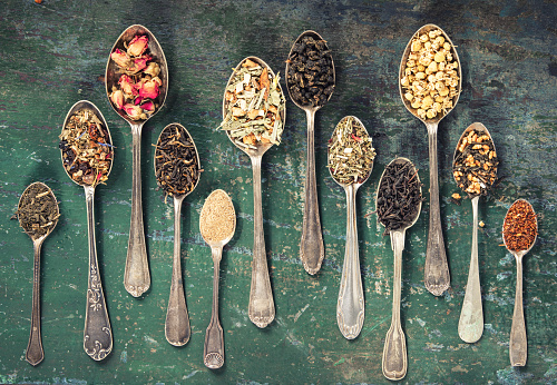 A collection of ornate vintage spoons, each cradling a different type of dried tea leaf, arrayed on a distressed green background.