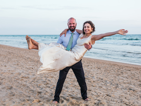 A joyful moment of a groom carrying his bride on a sandy beach with waves in the background.