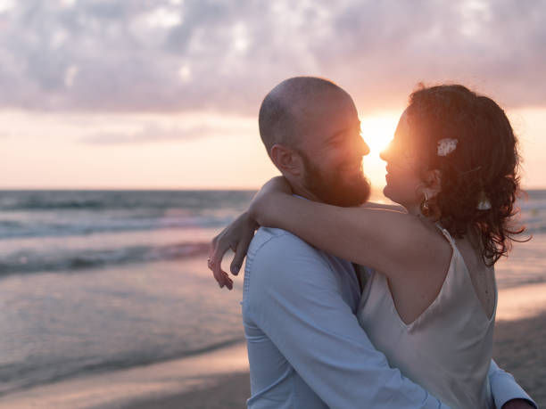 A couple embraces on a beach, the sun setting behind them casts a warm glow.