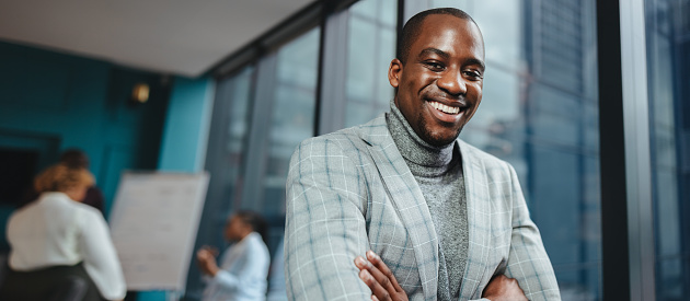 Confident African male entrepreneur stands in a boardroom full of people during a business meeting. He wears a suit, has arms crossed, and smiles at the camera, exuding professionalism and success.