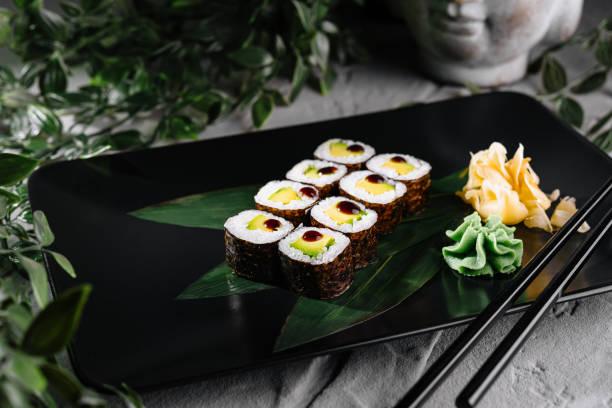 Sushi rolls garnished with wasabi and ginger on a modern black plate, botanical backdrop