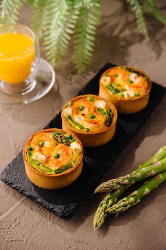Gourmet mini quiches with fresh asparagus on a slate plate, paired with orange juice and fern decoration