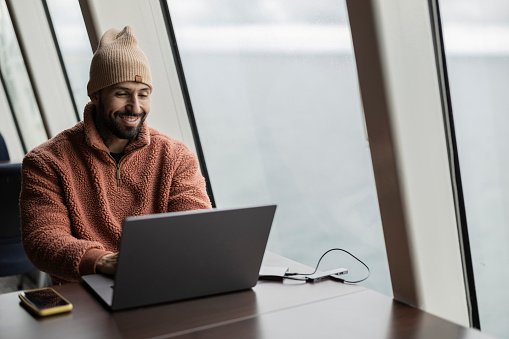 A smiling man in a beanie enjoys his work on a laptop, sitting comfortably in a modern coastal office.