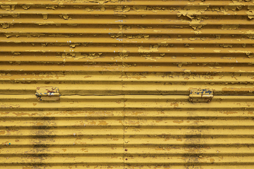 Fragment of an old metal wall. The corrugated steel sheets are connected with rivets and hinges. The yellow paint partially bulged and fell off. Background. Texture.