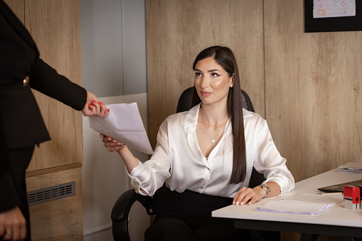 A female lawyer seated at her desk receives documents from a colleague, collaborating on a legal case.