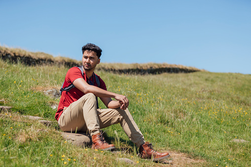 Full length wide side view of a young man sitting on the rocks wearing a red T-shirt and beige trousers.  He is looking at the view from the top of the hill on a summer's day at Hadrian's Wall, Northumberland.