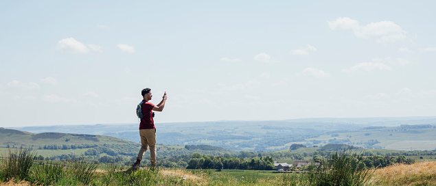 Full length rear view of a young man standing at the top of a hill in a red T-shirt and beige trousers. He is taking a photo using his smartphone of the view on a summer's day at Hadrian's Wall, Northumberland.