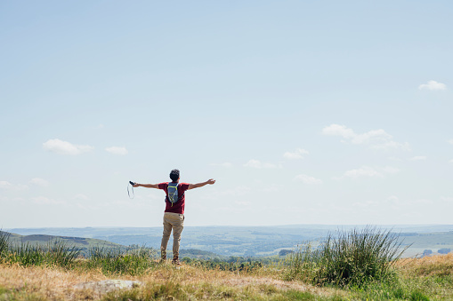 Full length rear wide view of a young man who has completed his hike to the top of a hill, he is wearing a red T-shirt, beige trousers and a backpack. He is standing with his arms spread wide and he is taking in the view on a summer's day at Hadrian's Wall, Northumberland.