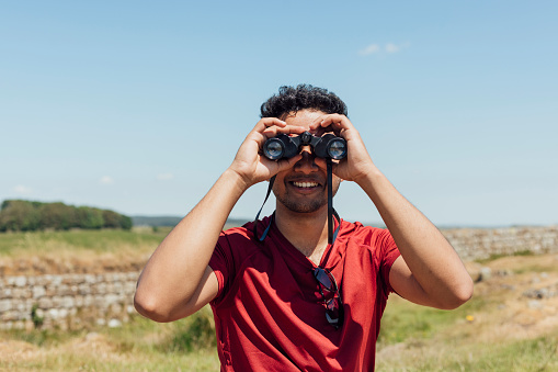 Waist up front view of a young male hiker walking along a footpath in a red T-shirt. He is using a pair of binoculars to look at the view on a summer's day at Hadrian's Wall, Northumberland.