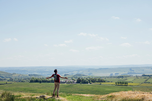 Full length rear wide view of a young man who has completed his hike to the top of a hill, he is wearing a red T-shirt, beige trousers and a backpack. He is standing with his arms spread wide and he is taking in the view on a summer's day at Hadrian's Wall, Northumberland.