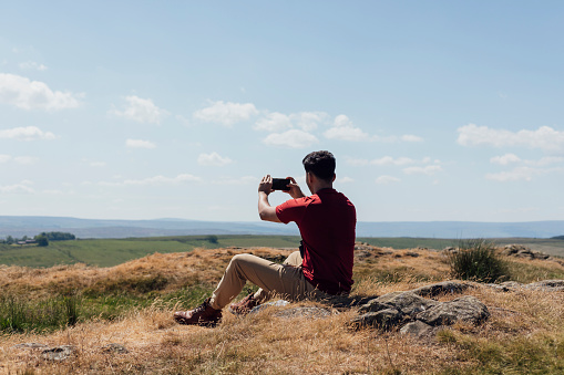 Full length rear view of a young man sitting on a rock in a red T-shirt and beige trousers. He is taking a photo using his smartphone of the view on a summer's day at Hadrian's Wall, Northumberland.