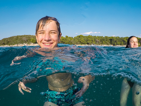 Happy teenage boy cooling off in the sea with his sister, heads above and bodies under water surface on a clear summer day, taken with wearable underwater camera